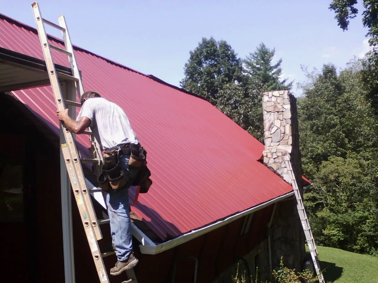 Roofer on a ladder beside a red metal roof and stone chimney — Soffit & Fascia Repair by Roofing Brothers in Hickory Hills